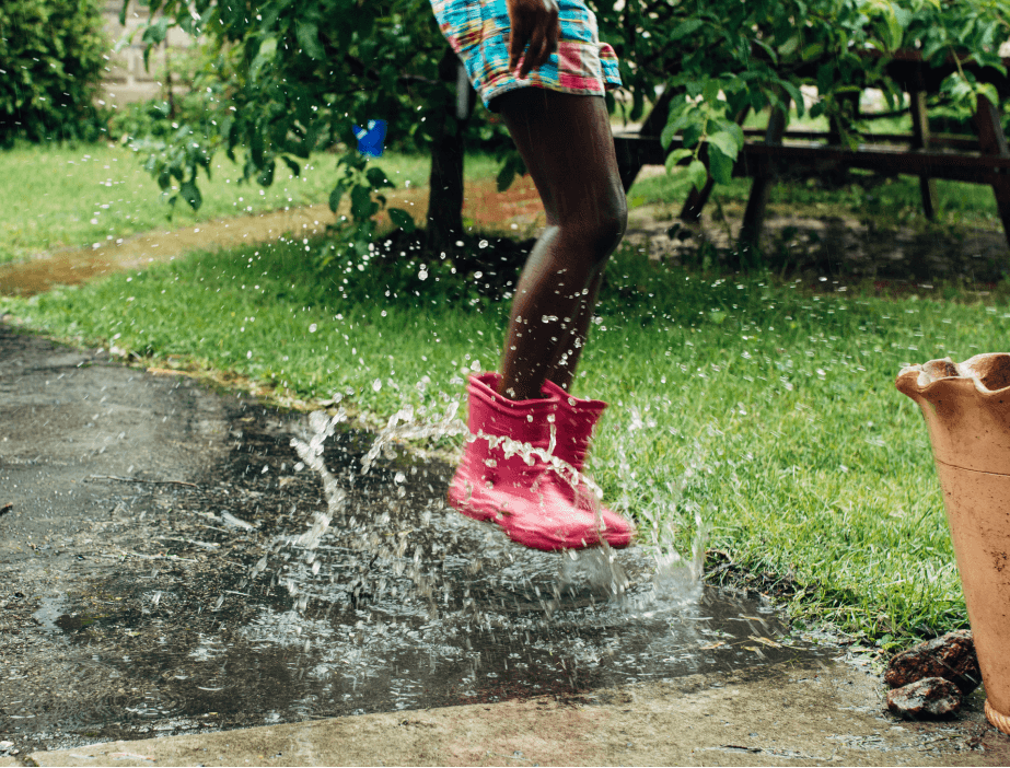 A child in rubber boots jumping in a puddle on the sidewalk.