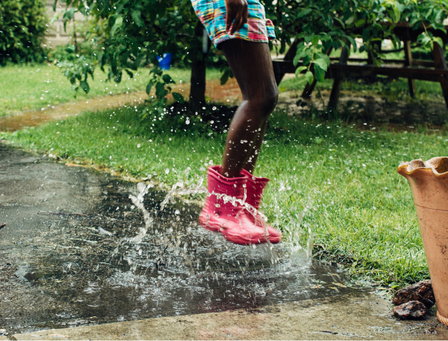 A child in rubber boots jumping in a puddle on the sidewalk.