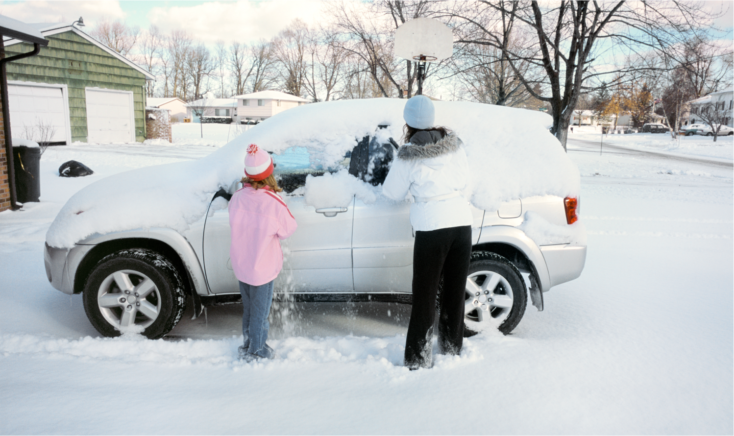 A mother and child wipe several inches of snow off their car in driveway.