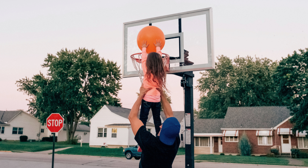 Man holding small girl up to basketball hoop so she can dunk.